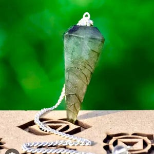 Op de afbeelding: Een gefacetteerde, doorschijnende kristallen pendel met een zilveren ketting en een heldere kristallen bol aan het einde. Het kristal is lichtgroen met donkere insluitsels. De pendel rust op een houten oppervlak met gesneden symbolen, tegen een vage groene achtergrond.