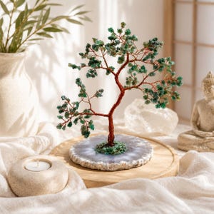 May include: A decorative wire tree sculpture featuring a twisted copper-coloured trunk and branches adorned with small, tumbled green gemstone leaves. The tree is mounted on a circular, translucent white agate slice base with a rough, natural edge.