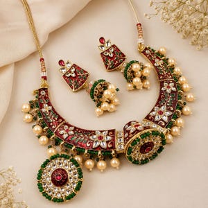 May include: A gold-toned necklace and earring set featuring deep red enamel with green leaf accents, clear stones, and cream-coloured pearl beads. The necklace has a large circular pendant, while the earrings are diamond-shaped with pearl drops.