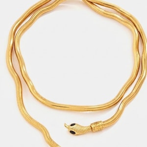 May include: A gold-colored snake necklace with a detailed snake head and tail clasp. The necklace is made of a flexible, textured material, and the snake head features black eye accents. The design is elegant and eye-catching.
