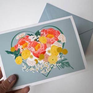 May include: A greeting card featuring a heart-shaped floral arrangement. The flowers are pink, yellow, and white, and the background is a light blue colour.