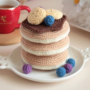 May include: A stack of crocheted pancakes with brown, beige, and white yarn. The pancakes are topped with crocheted blueberries and a beige crocheted circle. The pancakes sit on a white plate with decorative handles. A red mug is in the background.