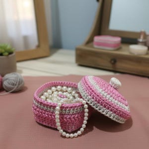 May include: A handmade pink crochet jewelry box with a matching lid, filled with white pearls. The box has a gray trim and a white knob on the lid. A strand of pearls drapes over the side. The box is on a pink surface.