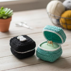 Puede incluir: Cajas de anillos de ganchillo, una negra con un lazo blanco y la otra turquesa, que revelan un anillo de oro. Las cajas están hechas a mano y se encuentran sobre una superficie de madera. Una pequeña planta suculenta y lana en el fondo.