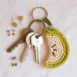 May include: A metal keyring holding two silver-toned keys and a semi-circular crochet charm shaped like a lime slice. The charm features a cream-colored center with brown embroidered seeds and a textured lime green border attached by a green ribbon.