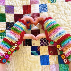 May include: A pair of colorful, crocheted fingerless gloves with a rainbow striped pattern. The gloves have red trim and are adorned with various colored buttons. The background is a patchwork quilt.