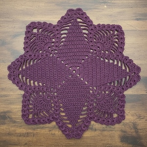 May include: A handmade, star-shaped crochet doily in a deep purple hue. The intricate design features a central square, radiating points, and a scalloped edge. The doily is crafted with a tight stitch, showcasing the detailed openwork pattern.