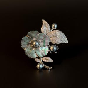 Puede incluir: Broche floral plateado con una gran flor iridiscente, perla central y detalles de diamantes. El diseño incluye hojas incrustadas con diamantes y acentos de perlas adicionales, sobre un fondo negro.