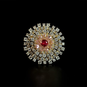 Puede incluir: Broche circular dorado con una gema roja central rodeada de cristales rosas y transparentes. El broche tiene un diseño de rayos de sol, con los cristales irradiando hacia afuera. El fondo es negro.