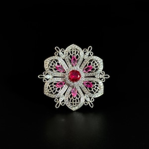 May include: A silver-toned flower-shaped brooch with a large, round, red gemstone center. The brooch features a detailed openwork design with smaller pink and clear gemstones, set against a black background.