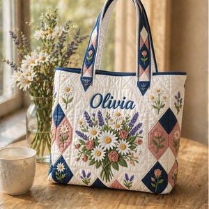 May include: A quilted tote bag with a floral design. The bag features a bouquet of daisies, roses, and lavender, with the name "Olivia" embroidered in blue. The bag has white, pink, and navy blue accents and a matching handle.