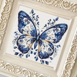 Può includere: Un'opera d'arte incorniciata con un design a farfalla blu e bianco. La farfalla presenta motivi intricati ed è circondata da accenti floreali. La cornice è color crema con dettagli ornati, creando un'estetica classica.