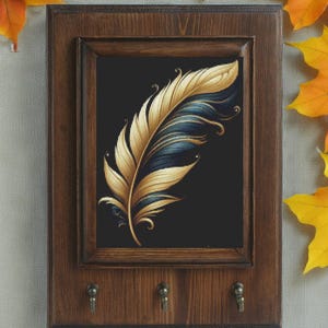 Può includere: Portachiavi in legno con un'immagine incorniciata di una piuma dorata e blu su sfondo nero. Il portachiavi ha tre ganci in metallo nella parte inferiore. Foglie autunnali sullo sfondo.