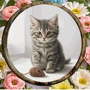 Könnte beinhalten: Ein Kreuzstichmuster eines grauen Tabby-Kätzchens mit braunen Augen, das sitzt und nach einer braunen Spielzeugmaus greift. Das Design ist in einem runden Rahmen in Holzoptik eingefasst, umgeben von rosa und weißen Blumen.