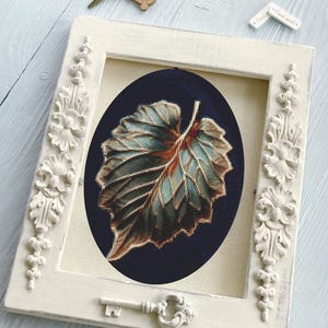 Può includere: Un'opera d'arte a punto croce incorniciata con un design dettagliato di foglie nei toni del verde acqua, marrone e arancione su uno sfondo blu scuro. La cornice bianca presenta dettagli floreali ornati e un accento chiave decorativo.