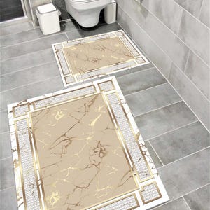Puede incluir: Dos alfombras rectangulares para baño con diseño de mármol. Las alfombras tienen un fondo beige con vetas doradas y un borde dorado. El borde exterior es blanco con un patrón texturizado. Las alfombras están colocadas sobre un suelo de baldosas grises.