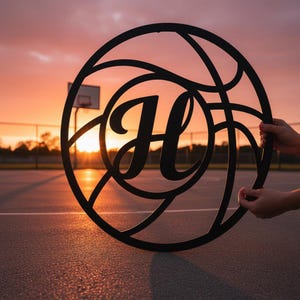 Könnte beinhalten: Schwarze Metall-Wandkunst im Basketball-Design mit dem Buchstaben "H" in der Mitte. Das Design ahmt das Muster eines Basketballs nach. Das Kunstwerk wird vor einem Sonnenuntergang auf einem Basketballplatz präsentiert.