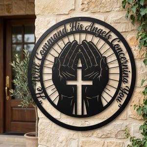 Könnte beinhalten: Schwarze Metall-Wandkunst mit einem Kreuz in gefalteten Händen, umgeben von dem Schriftzug "He Will Command His Angels Concerning You". Das kreisförmige Design ist an einer Steinmauer befestigt.