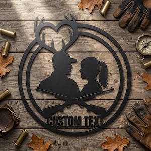 Könnte beinhalten: Ein rundes Wandbild aus schwarzem Metall mit den Silhouetten einer Person mit Baseballkappe und einer Person mit Pferdeschwanz unter einem herzförmigen Hirschgeweih-Design. Zwei gekreuzte Gewehre befinden sich unter den Figuren mit dem Text CUSTOM TEXT.