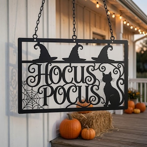 May include: A black metal hanging sign featuring the text Hocus Pocus in a decorative script font. The design includes three witch hats along the top, a spider web in the bottom left corner, and a silhouette of a cat on the right side, all suspended by chains.