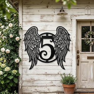 Könnte beinhalten: Schwarze Hausnummer 5 aus Metall mit Engelsflügeln. Die Flügel haben detaillierte Federn und ein Kreuzdesign. Die Nummer befindet sich in einem kreisförmigen Rahmen. Das Schild ist an einer weißen Holzwand befestigt.