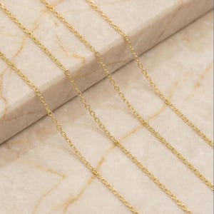 May include: Close-up of several delicate gold-colored chain necklaces. The chains are arranged diagonally across a light-colored, marble-like surface. The chains have small, evenly spaced links.