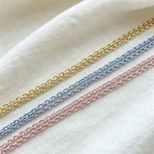 Pode incluir: Close-up de três delicados colares de corrente em ouro, azul claro e rosa. As correntes são feitas de pequenas ligações ovais espaçadas uniformemente. Os colares são exibidos em um fundo de tecido branco texturizado.