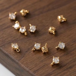 以下が含まれることがあります： クリアな正方形カットの宝石が付いた、小さな金色のジュエリーのクローズアップ。 各作品は、複雑なデザインと輝きを際立たせるために、暗い木製の表面に散らばっています。 ジュエリーはイヤリングまたはチャームのようです。