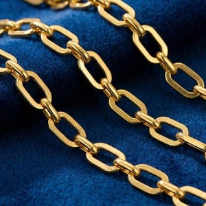 May include: Three gold-colored chain necklaces with oval links are arranged diagonally across a deep blue velvet surface. The chains have a polished finish, reflecting light. The background is a rich, dark blue, creating a contrast with the gold.