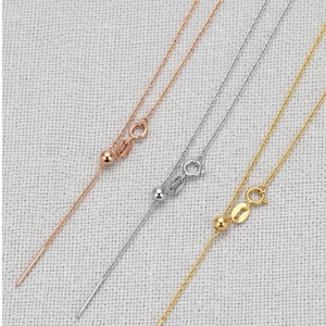 Puede incluir: Primer plano de tres delicados collares en oro rosa, plata y oro. Cada collar presenta una fina cadena con una pequeña bola y un colgante de barra. Los collares se exhiben sobre una superficie clara y texturizada.