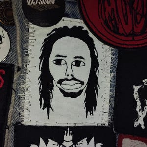Könnte beinhalten: Nahaufnahme einer Jeansjacke mit verschiedenen Aufnähern und Anstecknadeln. Ein zentraler Aufnäher zeigt eine Schwarz-Weiß-Illustration eines Männergesichts. Weitere Aufnäher zeigen Bandlogos und grafische Designs. Eine Anstecknadel mit dem Text "DJ Shadow" ist ebenfalls sichtbar.