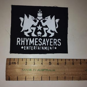 Puede incluir: Parche cuadrado negro con un gr&aacute;fico blanco de dos criaturas m&iacute;ticas, una corona y estrellas. El texto "RHYSAYERS ENTERTAINMENT" est&aacute; debajo. Una regla de madera con marcas en cm est&aacute; debajo.