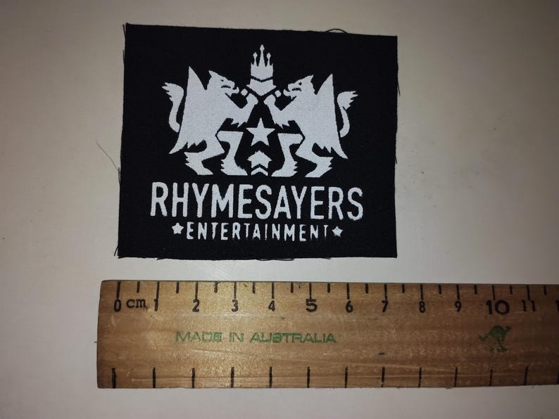 Puede incluir: Parche cuadrado negro con un gr&aacute;fico blanco de dos criaturas m&iacute;ticas, una corona y estrellas. El texto "RHYSAYERS ENTERTAINMENT" est&aacute; debajo. Una regla de madera con marcas en cm est&aacute; debajo.