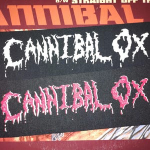 May include: Two black rectangular patches with the text "CANNIBAL OX" in a dripping, stylized font. One patch has white text, and the other has pink text. The patches are on a background with red and orange tones.