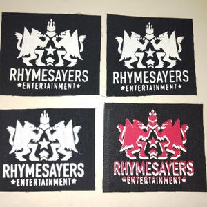 Puede incluir: Cuatro parches cuadrados negros con el texto "RHYSAYERS ENTERTAINMENT" en blanco. Los parches presentan un emblema blanco de dos criaturas aladas y una corona. Un parche tiene un emblema y texto rojos.