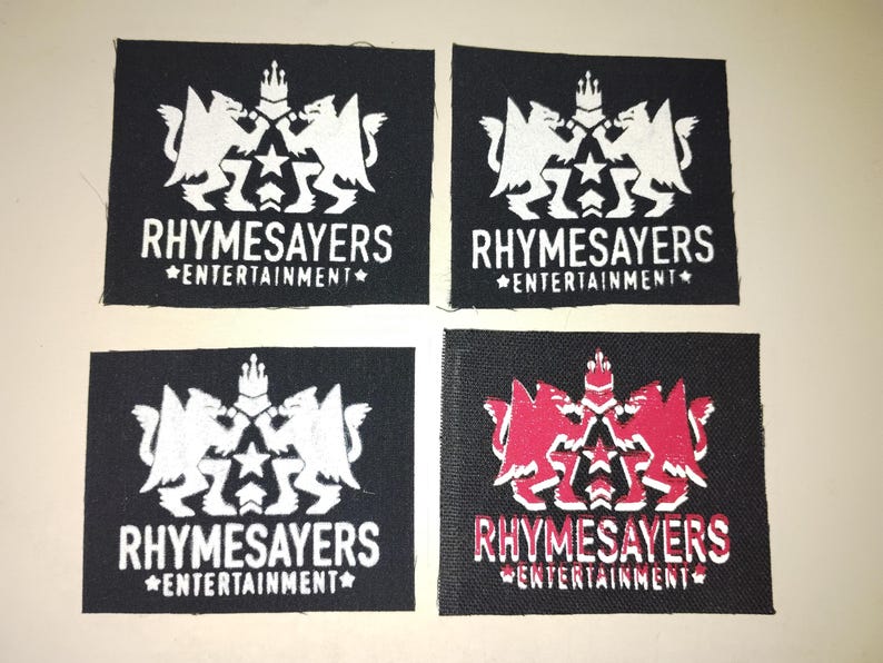 Puede incluir: Cuatro parches cuadrados negros con el texto "RHYSAYERS ENTERTAINMENT" en blanco. Los parches presentan un emblema blanco de dos criaturas aladas y una corona. Un parche tiene un emblema y texto rojos.
