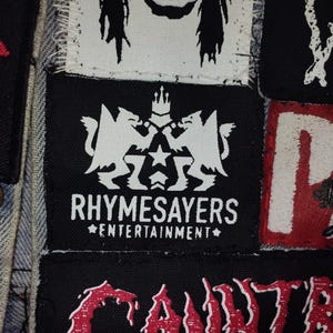 Puede incluir: Primer plano de una colecci&oacute;n de parches, principalmente en blanco y negro, con algunos detalles en rojo. El parche central presenta el texto "RHYSAMESAYERS ENTERTAINMENT" con un dise&ntilde;o de escudo. Otros parches muestran varios logotipos de bandas.
