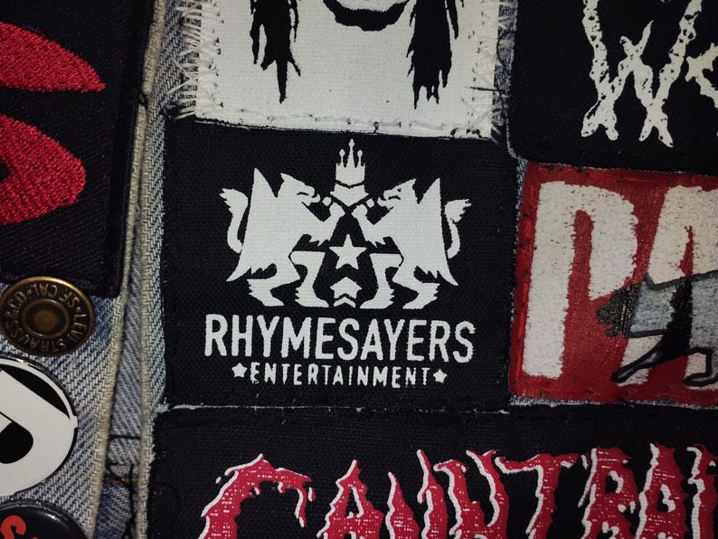 Puede incluir: Primer plano de una colecci&oacute;n de parches, principalmente en blanco y negro, con algunos detalles en rojo. El parche central presenta el texto "RHYSAMESAYERS ENTERTAINMENT" con un dise&ntilde;o de escudo. Otros parches muestran varios logotipos de bandas.