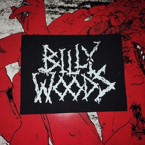 May include: A black rectangular patch with the white text "BILLY WOODS" in a stylized, jagged font. The patch is set against a red and white background featuring a wolf and a figure.