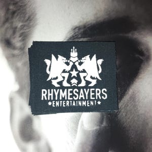May include: A black rectangular patch with the white text "RHYSAYERS ENTERTAINMENT" and a white emblem featuring two mythical creatures, a star, and a crown. The patch is stacked on top of other patches.