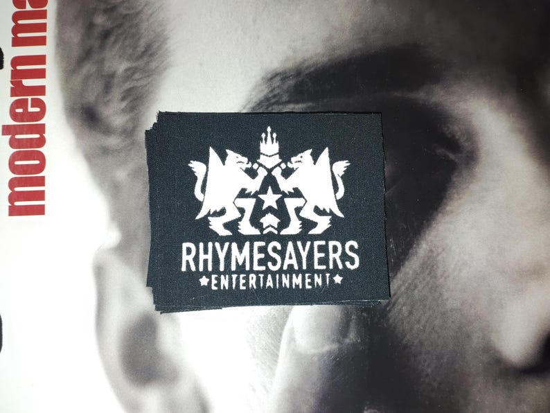 Puede incluir: Un parche rectangular negro con el texto blanco "RHYSAYERS ENTERTAINMENT" y un emblema blanco con dos criaturas m&iacute;ticas, una estrella y una corona. El parche est&aacute; apilado encima de otros parches.