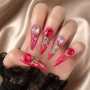Op de afbeelding: Een hand met vijf lange, puntige kunstnagels. Elke nagel is felroze geverfd met een glittertextuur en versierd met grote, kleurrijke edelstenen en goudkleurige versieringen. De edelstenen hebben verschillende vormen en kleuren, waaronder roze, helder en rood.