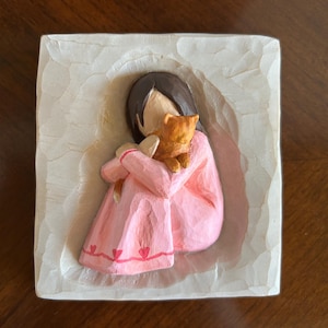 Puede incluir: Escultura en relieve cuadrada y blanquecina tallada. Representa una figura con un vestido rosa abrazando a un pequeño gato naranja. El vestido tiene un diseño de corazón rojo. La figura tiene cabello castaño y sostiene al gato cerca.