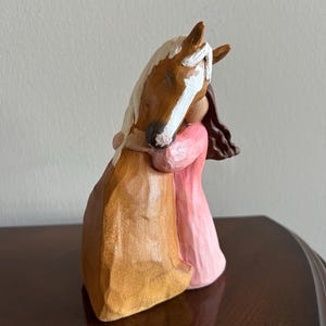 May include: A wooden sculpture of a horse embracing a person. The horse is brown with a white blaze and mane. The person is wearing a pink dress and has brown hair. The sculpture is on a dark brown surface.