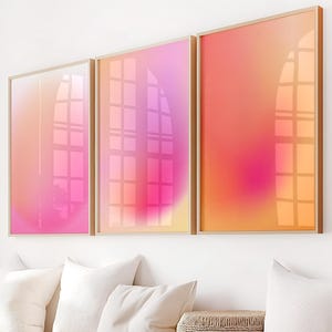 May include: Three rectangular wall art prints in light wood frames featuring a gradient of pink, orange, and yellow colors. The artwork displays a soft, blurred color transition with a subtle reflection of a window grid visible on the glass surface.