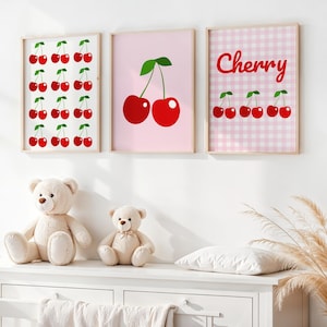 May include: Three framed art prints with cherry designs hang above a white dresser. The prints feature red cherries on white, pink, and a pink gingham background. Two teddy bears sit on the dresser. A decorative plant is to the right.