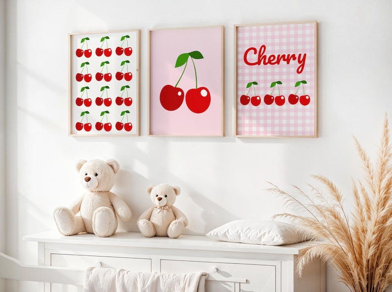 May include: Three framed art prints with cherry designs hang above a white dresser. The prints feature red cherries on white, pink, and a pink gingham background. Two teddy bears sit on the dresser. A decorative plant is to the right.
