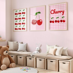 May include: Three framed cherry-themed art prints in a child's room. One print features a grid of cherries, another shows two cherries, and the third says "Cherry" with cherry illustrations on a pink gingham background. A teddy bear and storage bench are in the foreground.