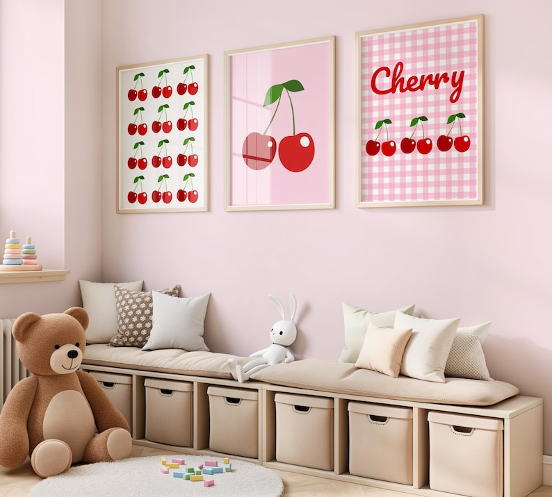 May include: Three framed cherry-themed art prints in a child's room. One print features a grid of cherries, another shows two cherries, and the third says "Cherry" with cherry illustrations on a pink gingham background. A teddy bear and storage bench are in the foreground.