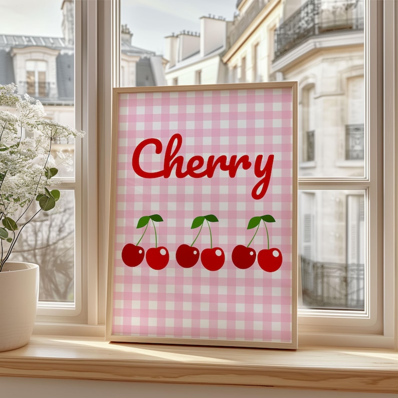 May include: A framed print featuring the word "Cherry" in red script above three pairs of red cherries with green leaves. The background is a pink and white gingham pattern. The frame is a light wood color.